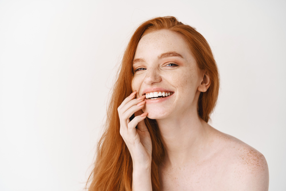 close up of happy redhead woman with pale perfect skin laughing and showing white teeth standing naked on studio wall
