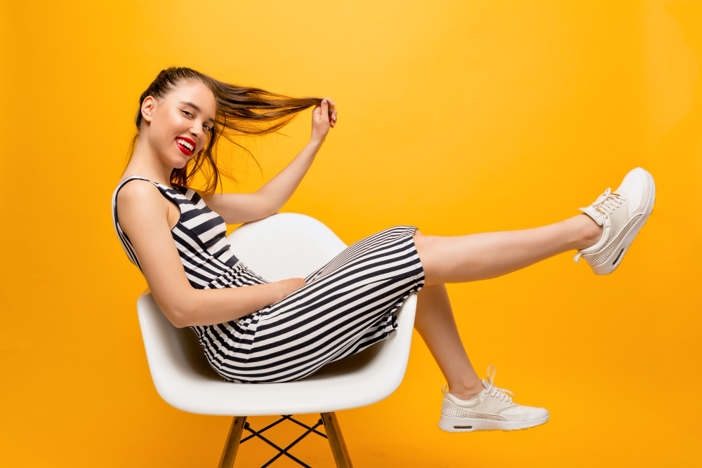 full lenght photo of stylish happy woman with red lips hair dressed fitting dress and white sneakers sitting in the chair and plays with her hair over yellow wall place for